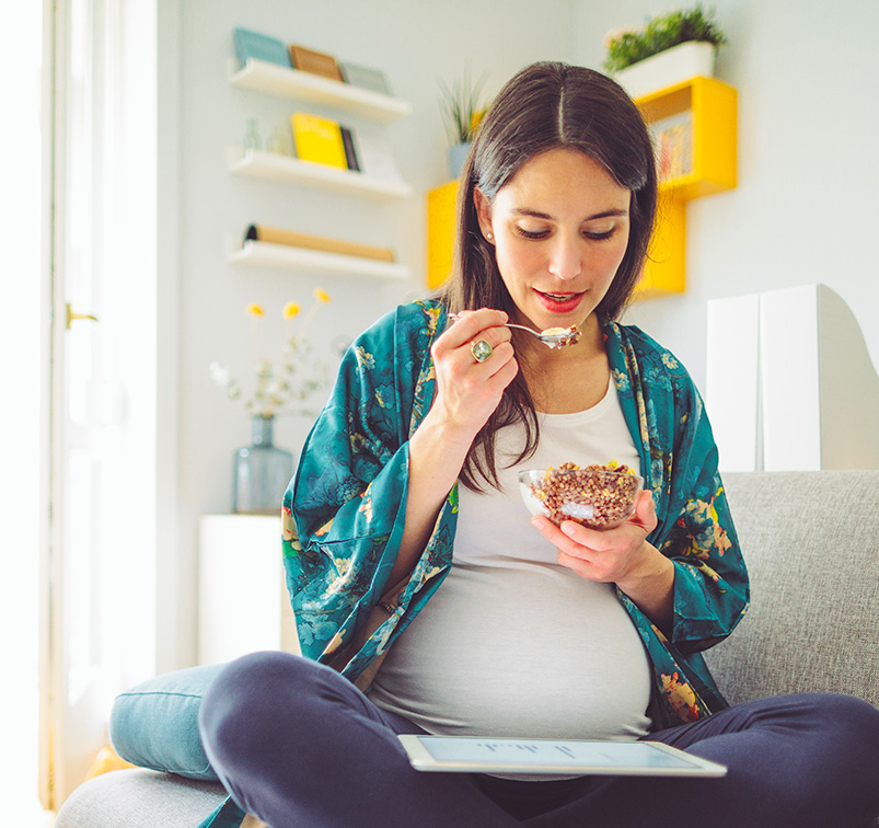 Junge, schwangere Frau sitzt auf einem Sofa mit einem Pad-Computer auf dem Schoß und einer Schüssel mit Körnern und weißer Creme zum Essen in den Händen.